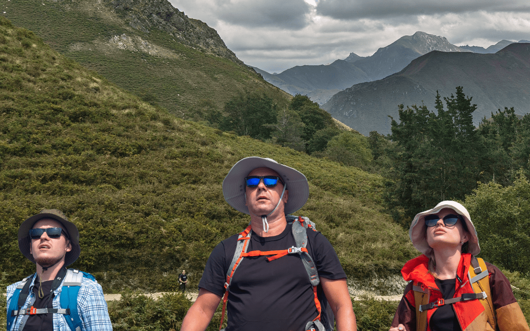 Tres peregrinos con equipamiento de senderismo recorren un tramo montañoso y verde del Camino de Oviedo a Covadonga, en la etapa desde el Alto del Espinadal hasta Puente Miera, con bosques frondosos y cumbres asturianas al fondo