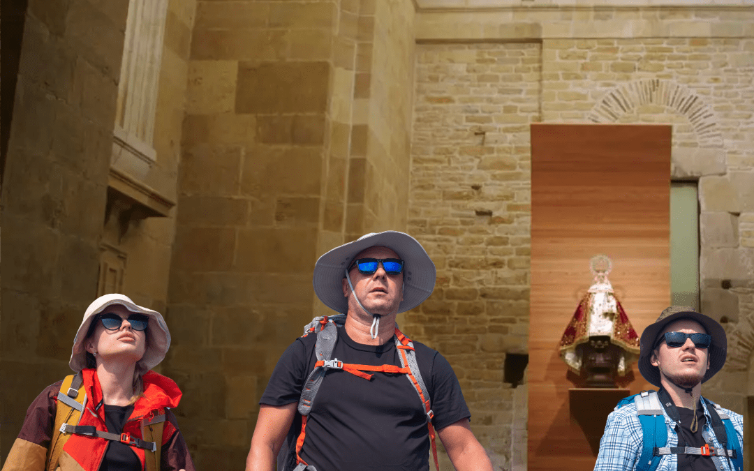 Tres peregrinos equipados con mochilas y bastones de senderismo en el interior de la Capilla de la Virgen de Covadonga, origen del Camino de Oviedo a Covadonga, con muros de piedra histórica y la imagen de la Santina al fondo.