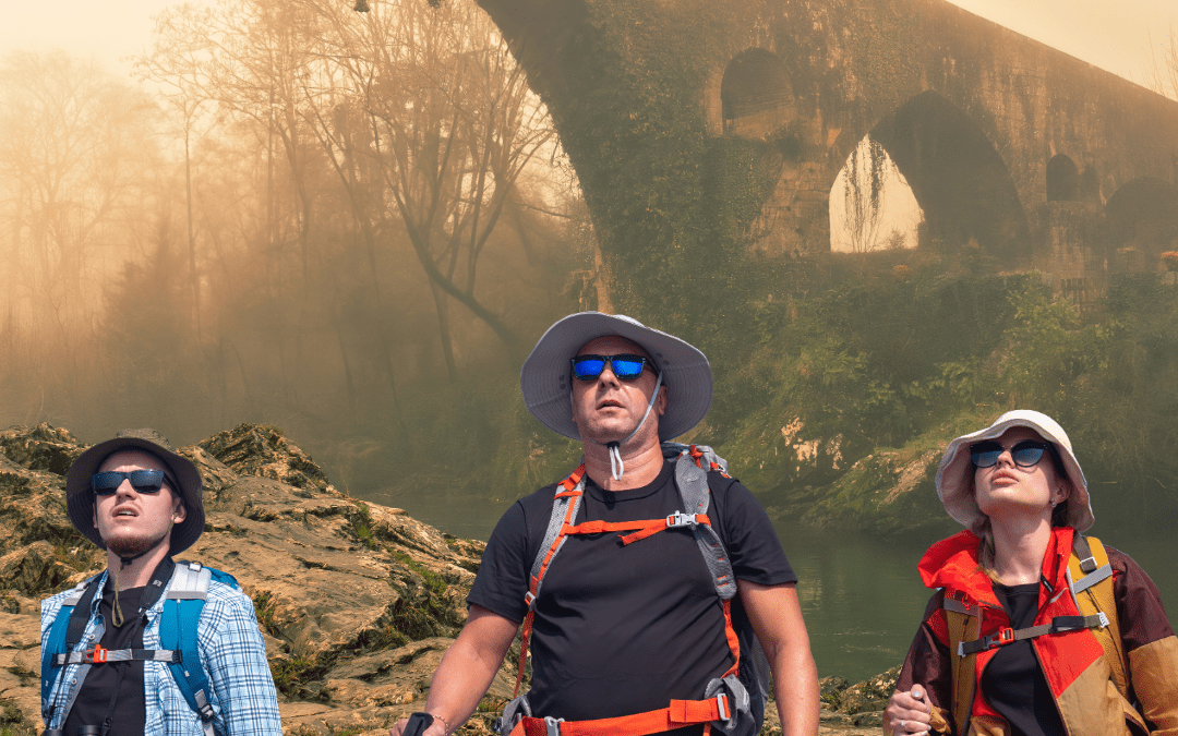 Tres senderistas con mochilas, sombreros y bastones de trekking están en primer plano sobre un terreno rocoso junto a un río, mientras al fondo se ve el Puente Romanos , un antiguo puente de piedra cubierto de vegetación y envuelto en niebla dorada, con una cruz colgando bajo uno de sus arcos en un entorno boscoso.